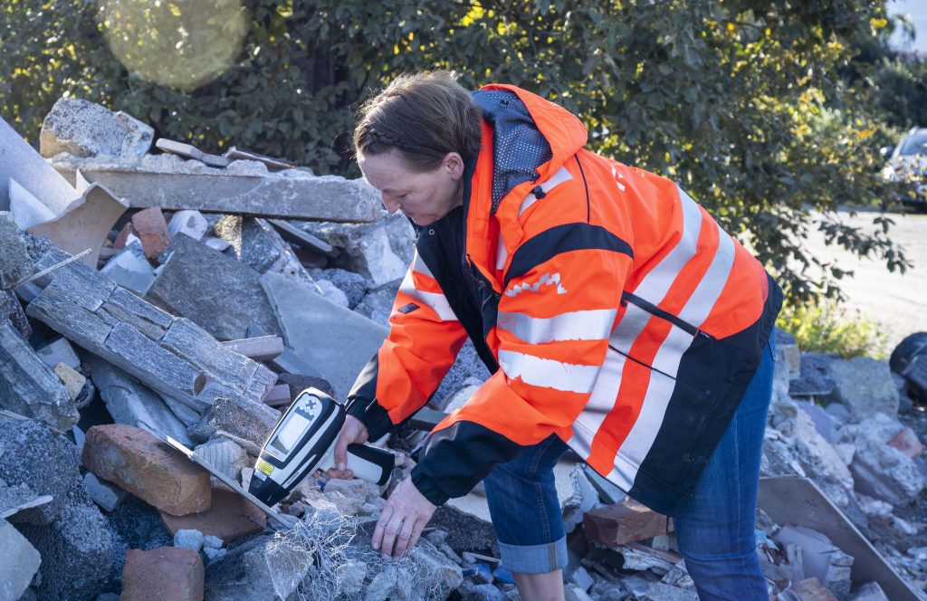 Bilde av Lise som måler farlig avfall med et måleinstrument som ser ut som en vannpistol. Haugen hun måler er fylt med stein, murstein, tegl, treplater og Lecablokker. Lise har på seg en knalloransje arbeidsjakke det står Retura på.